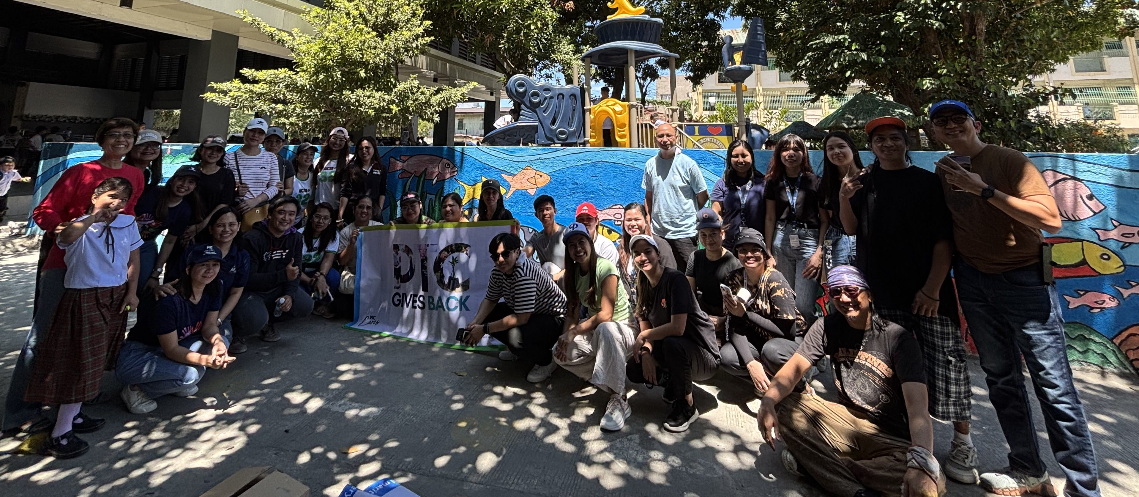 A large group of volunteers and students posing together in front of the completed long mural wall.