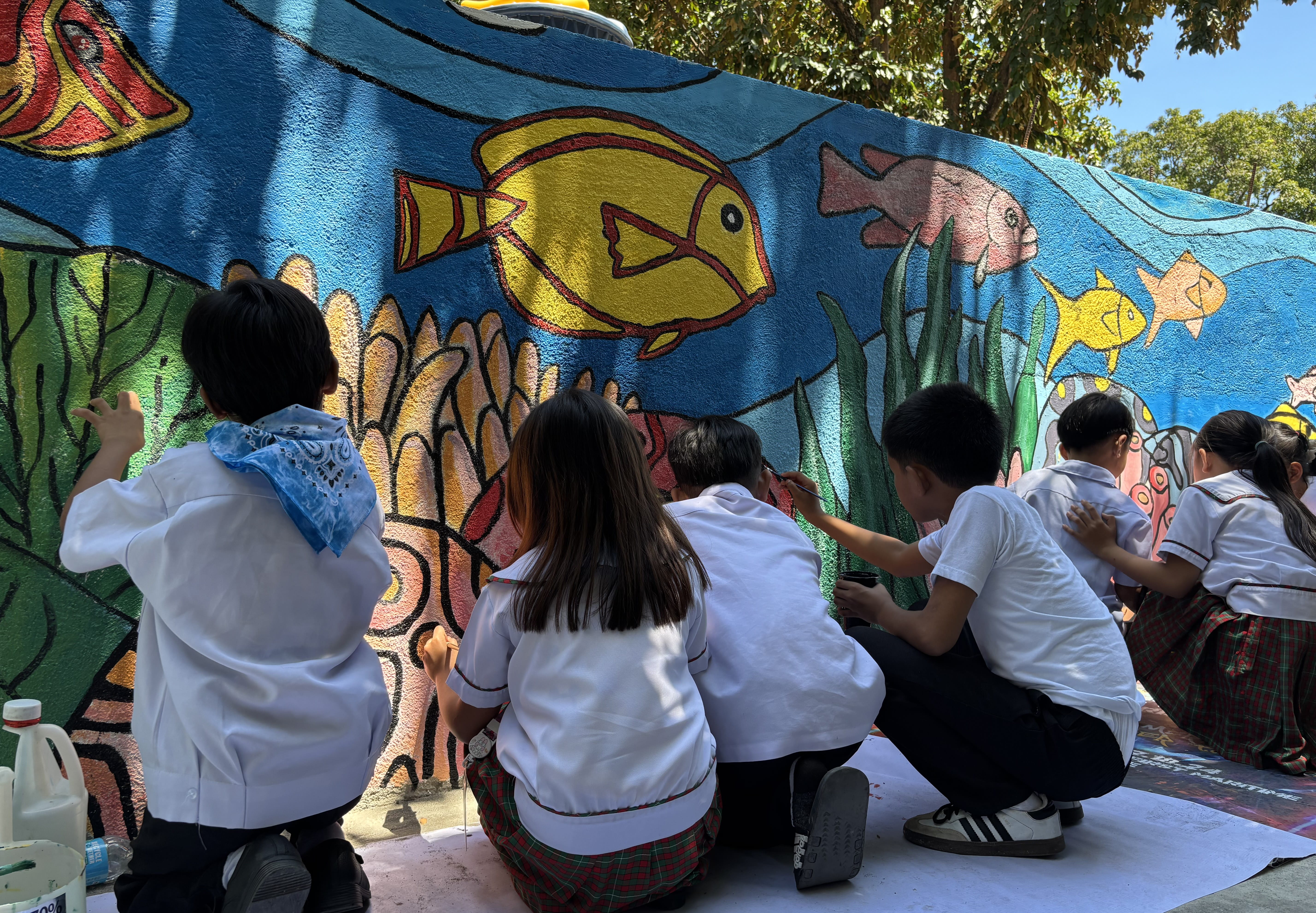 Students in school uniforms kneeling and painting colorful coral and fish on the lower section of the wall.